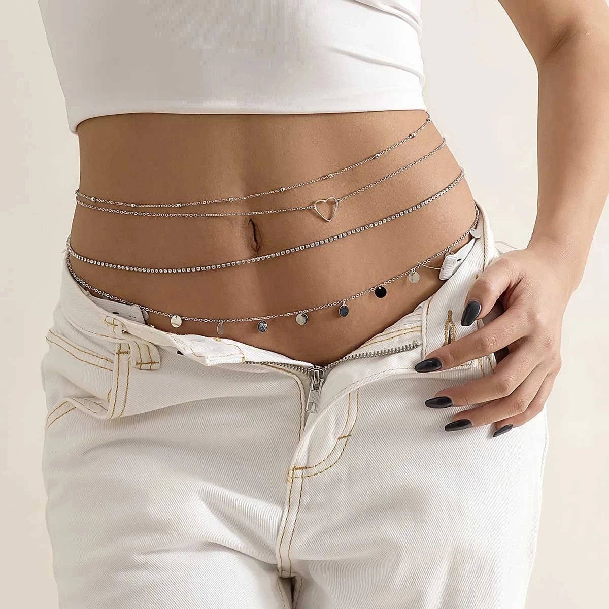 Woman wearing layered silver waist chains with heart charm and disc pendants over white jeans