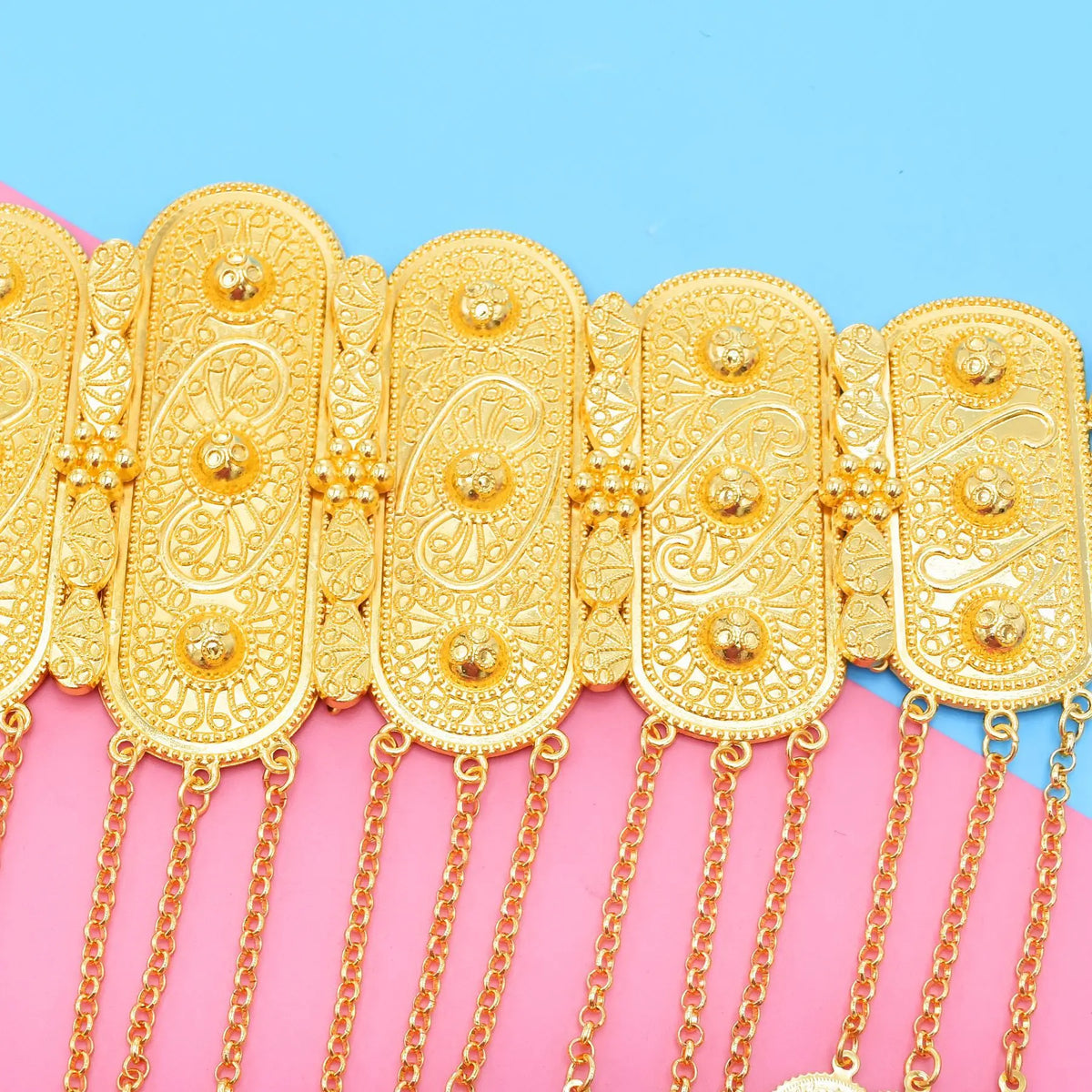 Intricately designed gold metal belt with ornate patterns and hanging chains on pastel background