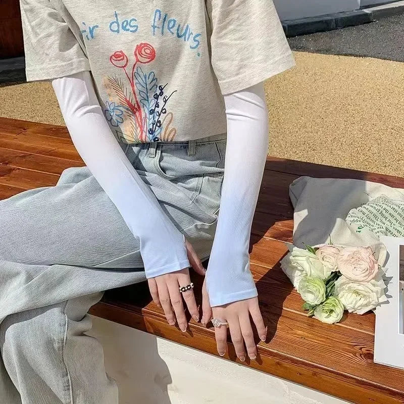 Woman wearing light grey t-shirt with floral print, long white arm sleeves, and light jeans, sitting on a wooden bench beside white roses.