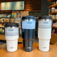 Three TYESO vacuum insulated stainless steel bottles in white and black on a wooden table in a cafe.