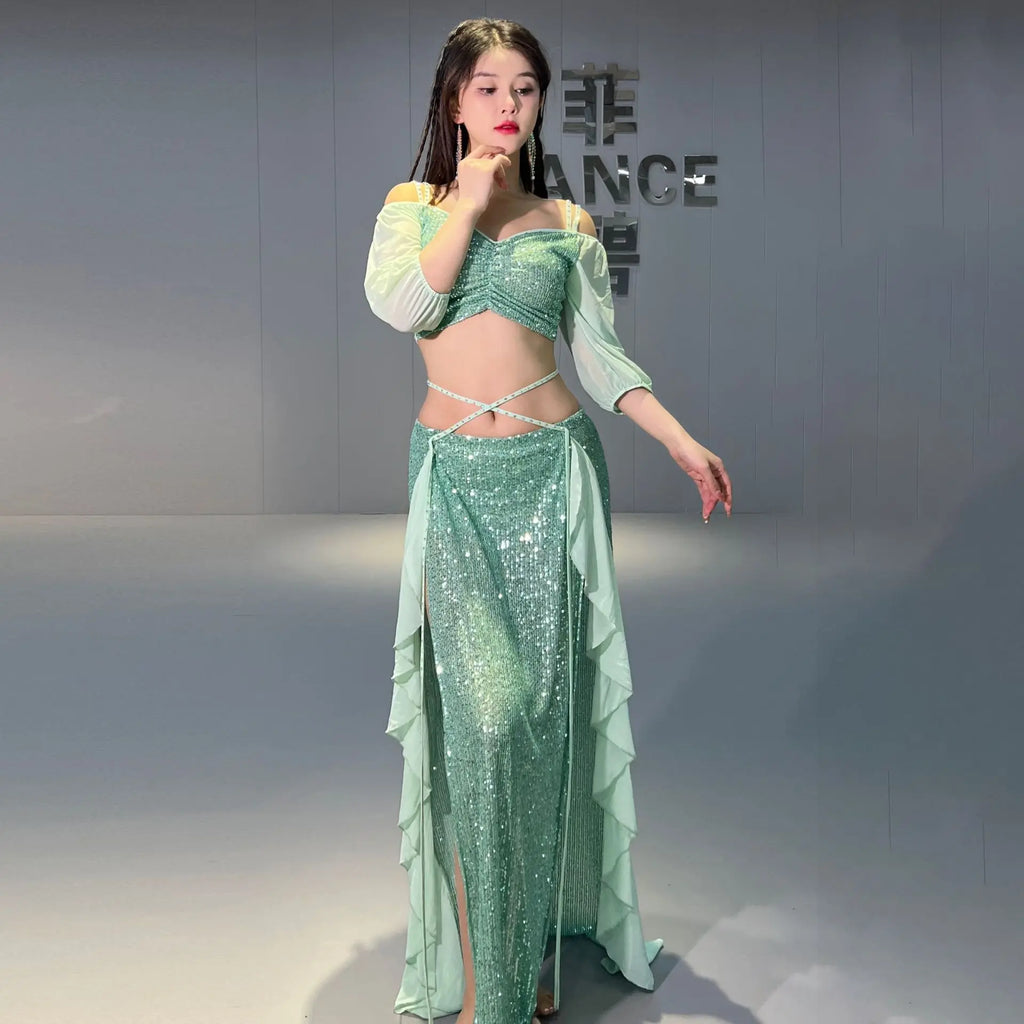 Woman in green sequin belly dance costume with sheer sleeves and ruffled skirt standing indoors