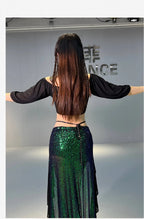 Woman in a black off-shoulder top and green sequin skirt standing with arms outstretched