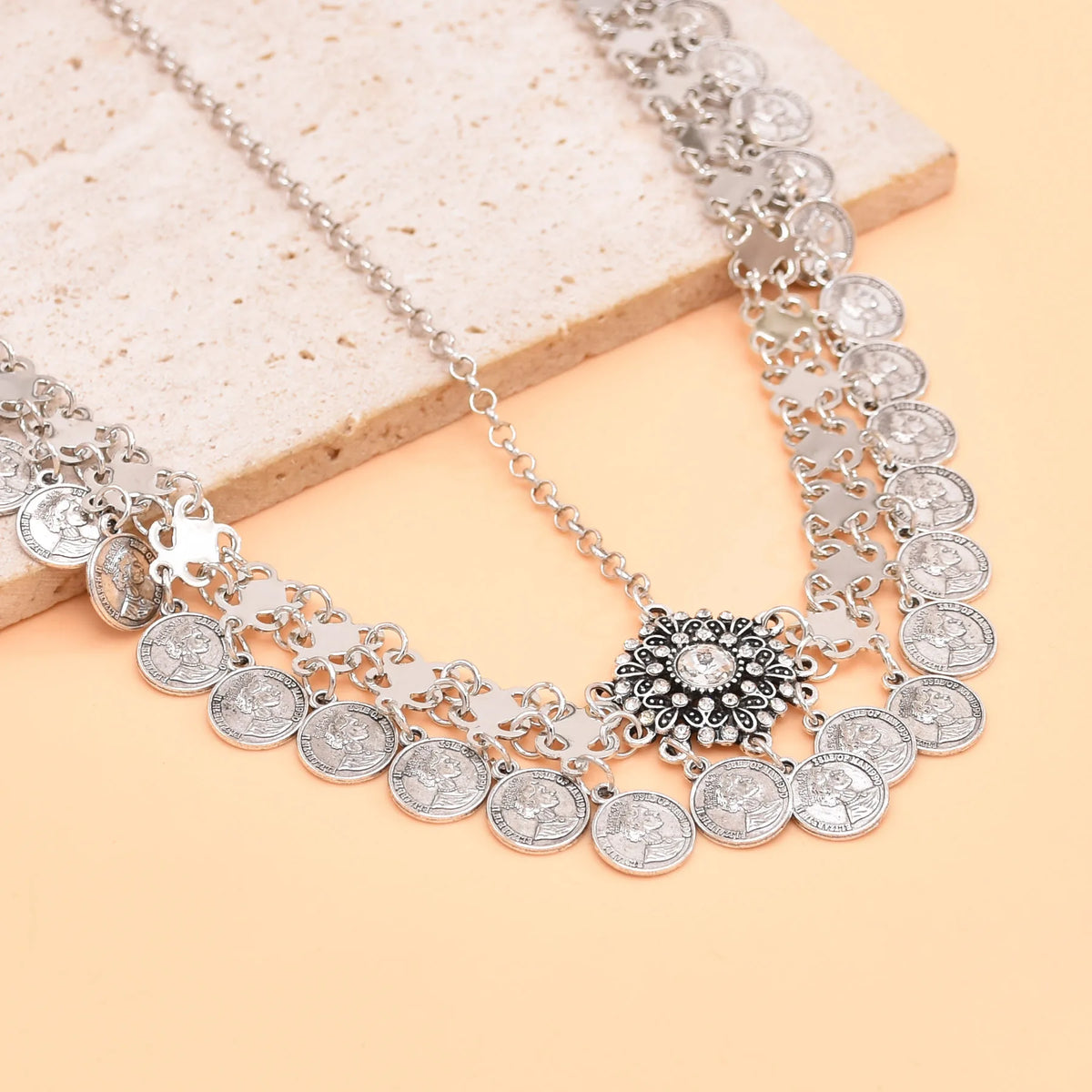 Silver coin necklace with ornate floral pendant on a beige stone background