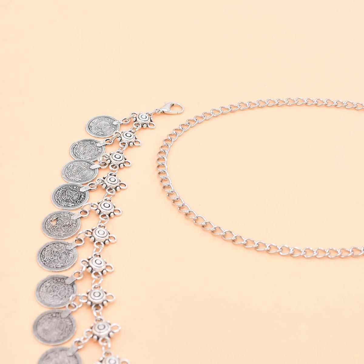 Silver coin charm anklet with intricate detailing on a light peach background