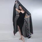 Woman in black dress with sheer black shawl adorned with colorful pompom trim, posing indoors.