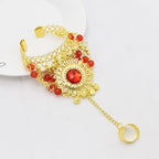 Gold hand chain bracelet with red beads and central red gemstone, attached ring design.