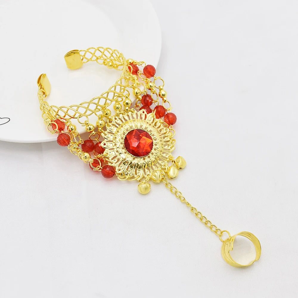 Gold hand chain bracelet with red beads and central red gemstone, attached ring design.